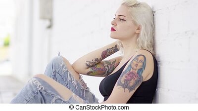 Sexy sultry young woman with a vampire tattoo on her arm sitting leaning  against a white outdoor wall turning to stare at the | CanStock