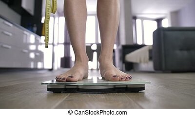 Disappointing weight results on bathroom scale. Young woman in lingerie  with measuring tape in hands checking her weight. | CanStock