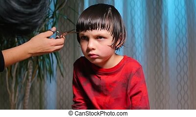 A serious child on a haircut by a hairdresser. a boy in red clothes is  scissored by cutting bangs on his head. | CanStock